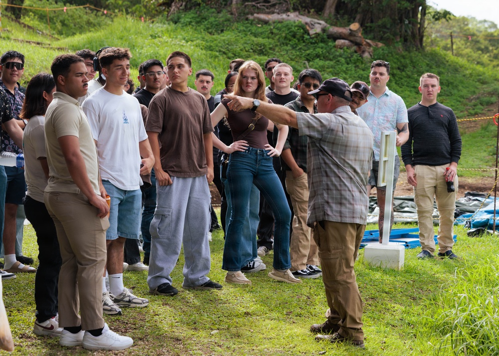 An instructor or guide is addressing a large group of young people outdoors on a grassy hillside. The person facing away from the camera appears to be pointing and explaining something, possibly conducting a field demonstration or educational tour. The group is attentively listening in an informal outdoor setting with lush green vegetation in the background.