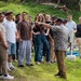 An instructor or guide is addressing a large group of young people outdoors on a grassy hillside. The person facing away from the camera appears to be pointing and explaining something, possibly conducting a field demonstration or educational tour. The group is attentively listening in an informal outdoor setting with lush green vegetation in the background.