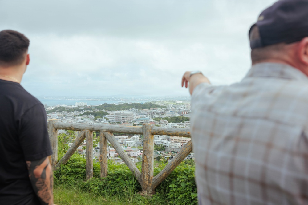 Two individuals are standing at an elevated overlook point, gazing at a coastal city spread out below them. One person on the right is pointing toward the cityscape or distant coastline, suggesting they are identifying a specific landmark or location. A rustic wooden log railing marks the edge of the viewpoint.