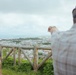 Two individuals are standing at an elevated overlook point, gazing at a coastal city spread out below them. One person on the right is pointing toward the cityscape or distant coastline, suggesting they are identifying a specific landmark or location. A rustic wooden log railing marks the edge of the viewpoint.