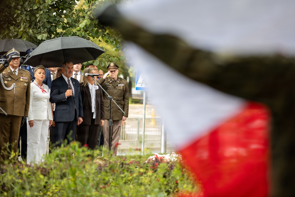 A formal outdoor ceremony is taking place, likely a military or state commemoration event. Several individuals including military officers in olive/khaki uniforms with medals and civilians in formal attire are gathered, with one officer rendering a salute. A red-and-white flag (consistent with Polish national colors) is visible in the foreground, partially blurred.