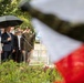 A formal outdoor ceremony is taking place, likely a military or state commemoration event. Several individuals including military officers in olive/khaki uniforms with medals and civilians in formal attire are gathered, with one officer rendering a salute. A red-and-white flag (consistent with Polish national colors) is visible in the foreground, partially blurred.