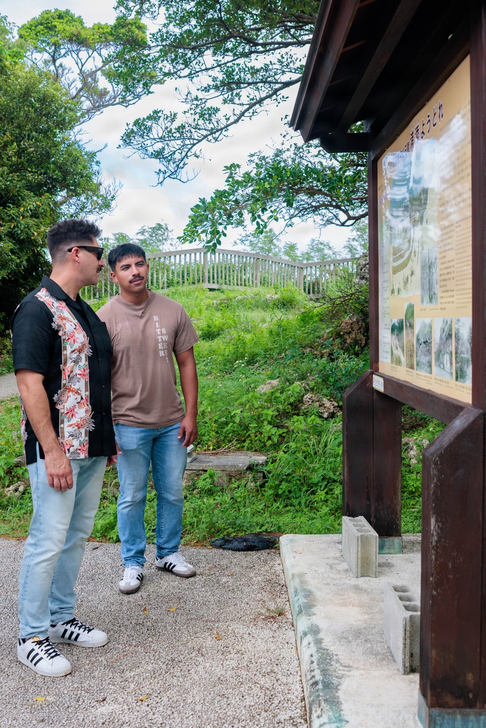 Two men are standing at an outdoor informational sign board with Japanese text and historical photographs, appearing to read or discuss its contents. The setting appears to be a historical or cultural site, likely in Okinawa, Japan, based on the Japanese signage and architectural style of the display structure. The surrounding environment features lush green vegetation, trees, and a wooden fence in the background.