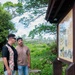 Two men are standing at an outdoor informational sign board with Japanese text and historical photographs, appearing to read or discuss its contents. The setting appears to be a historical or cultural site, likely in Okinawa, Japan, based on the Japanese signage and architectural style of the display structure. The surrounding environment features lush green vegetation, trees, and a wooden fence in the background.