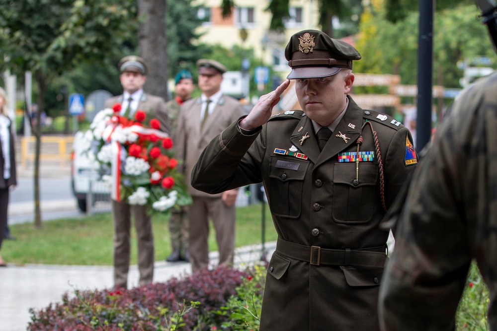 A U.S. Army officer in dress uniform renders a salute during what appears to be a formal memorial or wreath-laying ceremony. In the background, several individuals in military or formal attire stand at attention near a red-and-white floral wreath, consistent with Polish national colors. The setting appears to be an outdoor public space, likely in Poland.