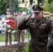 A U.S. Army officer in dress uniform renders a salute during what appears to be a formal memorial or wreath-laying ceremony. In the background, several individuals in military or formal attire stand at attention near a red-and-white floral wreath, consistent with Polish national colors. The setting appears to be an outdoor public space, likely in Poland.