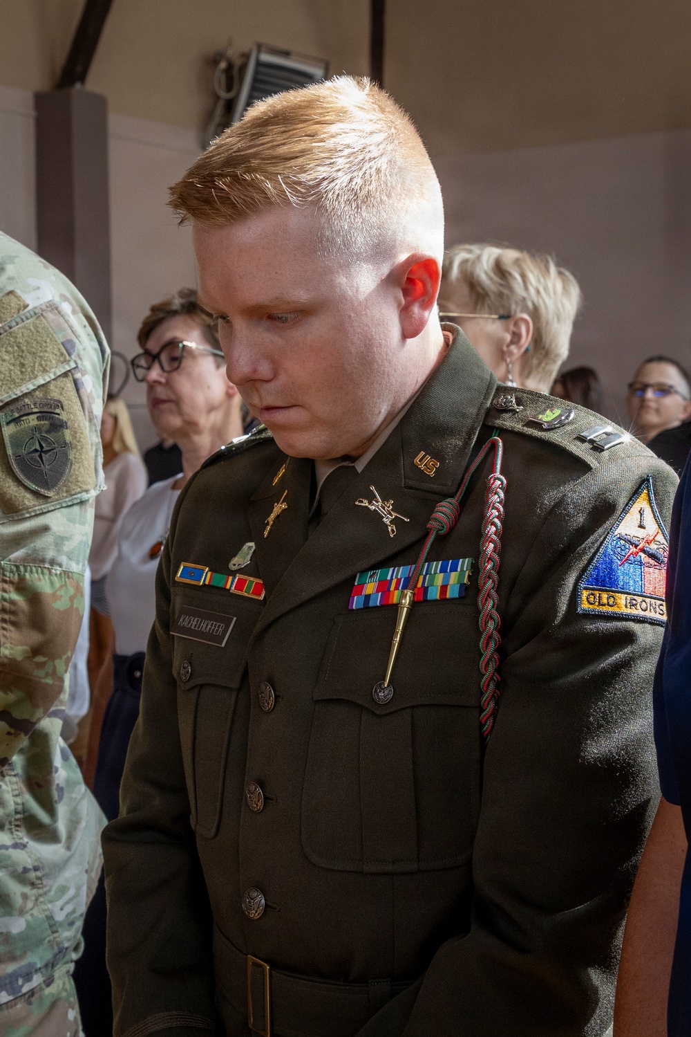 A U.S. Army soldier in Army Service Uniform (ASU) is standing with head bowed, possibly during a ceremony or prayer. The soldier wears multiple service ribbons, a unit crest, and a shoulder sleeve insignia reading 'OLD IRONS' associated with the 1st Armored Division. A name tape reading 'KACHELHOFFER' is visible on the uniform.