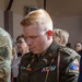 A U.S. Army soldier in Army Service Uniform (ASU) is standing with head bowed, possibly during a ceremony or prayer. The soldier wears multiple service ribbons, a unit crest, and a shoulder sleeve insignia reading 'OLD IRONS' associated with the 1st Armored Division. A name tape reading 'KACHELHOFFER' is visible on the uniform.