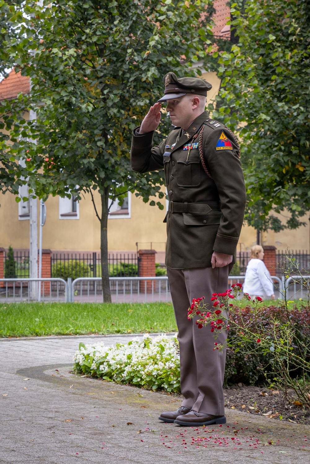 A U.S. Army officer in dress green uniform is standing at attention and rendering a salute, likely during a formal ceremony or memorial event. The soldier wears the 2nd Armored Division 'Old Ironsides' shoulder patch and multiple service ribbons. The setting appears to be an outdoor public square or memorial garden in a European town.