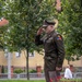 A U.S. Army officer in dress green uniform is standing at attention and rendering a salute, likely during a formal ceremony or memorial event. The soldier wears the 2nd Armored Division 'Old Ironsides' shoulder patch and multiple service ribbons. The setting appears to be an outdoor public square or memorial garden in a European town.