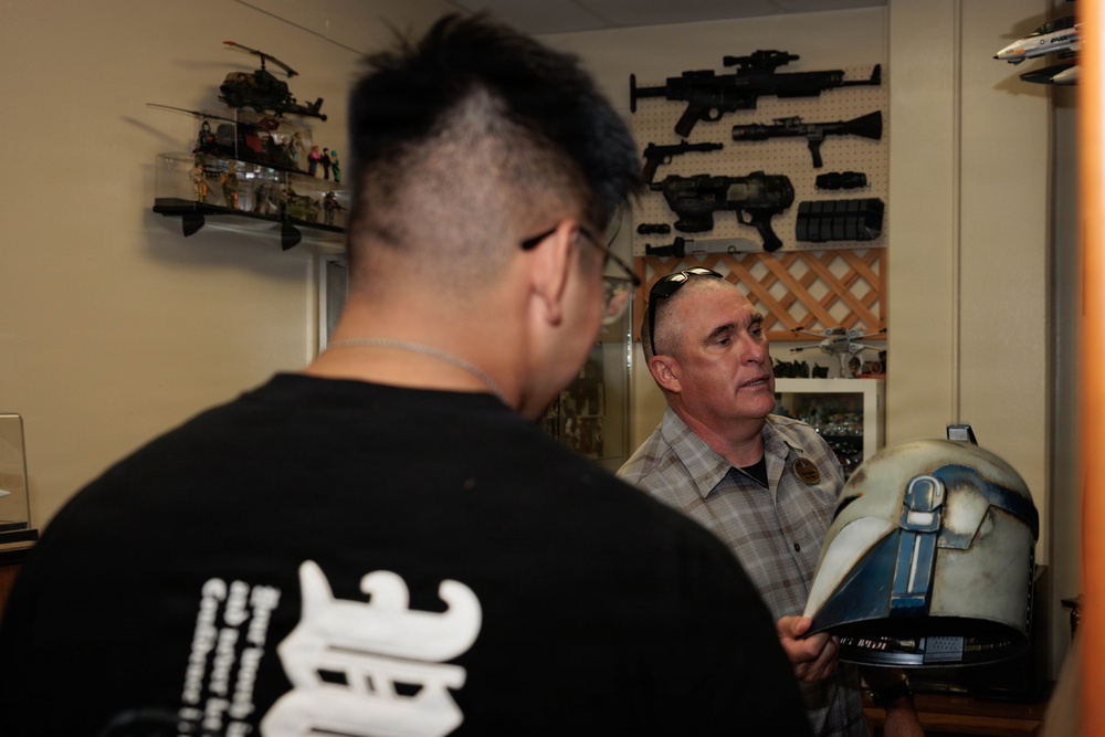 Two men are in a collector's room examining what appears to be a Star Wars-style helmet or prop. One man faces away from the camera wearing a black graphic t-shirt, while the other, an older man in a plaid shirt, holds and examines the helmet. The room is filled with collectibles including replica weapons mounted on a pegboard wall and action figures on shelves.