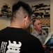 Two men are in a collector's room examining what appears to be a Star Wars-style helmet or prop. One man faces away from the camera wearing a black graphic t-shirt, while the other, an older man in a plaid shirt, holds and examines the helmet. The room is filled with collectibles including replica weapons mounted on a pegboard wall and action figures on shelves.