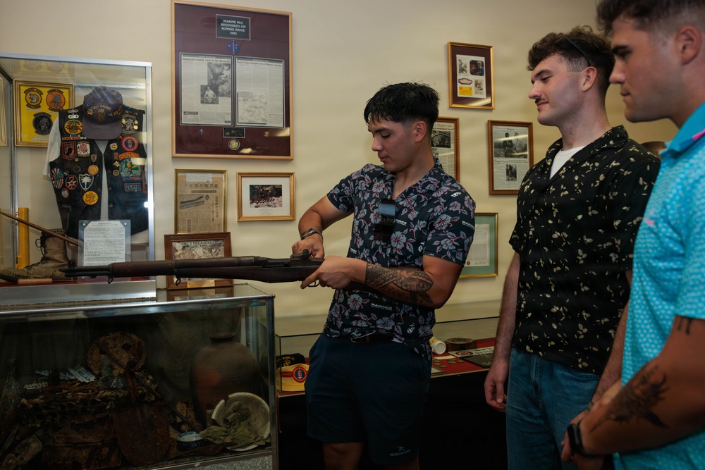 Three young men are examining what appears to be a historical rifle or long gun inside a military museum or memorial exhibit. The central figure, who has tattoos and is wearing a floral shirt, is holding and inspecting the firearm while the other two observe. The room is filled with military memorabilia including patches, framed documents, newspaper clippings, and display cases.