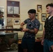 Three young men are examining what appears to be a historical rifle or long gun inside a military museum or memorial exhibit. The central figure, who has tattoos and is wearing a floral shirt, is holding and inspecting the firearm while the other two observe. The room is filled with military memorabilia including patches, framed documents, newspaper clippings, and display cases.
