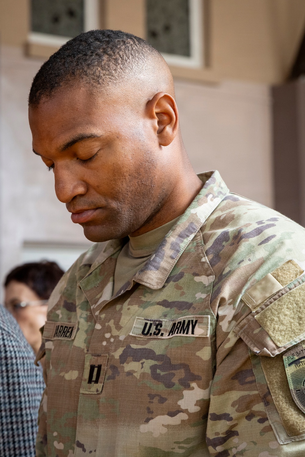 A U.S. Army soldier is photographed in close-up with head bowed downward, wearing an OCP (Operational Camouflage Pattern) uniform. The soldier's name tape reads partially as 'ARREN' and the branch tape reads 'U.S. ARMY,' with a rank insignia visible on the chest and a unit patch on the right shoulder. The background is softly blurred, showing a building exterior and another partially visible individual.
