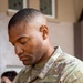 A U.S. Army soldier is photographed in close-up with head bowed downward, wearing an OCP (Operational Camouflage Pattern) uniform. The soldier's name tape reads partially as 'ARREN' and the branch tape reads 'U.S. ARMY,' with a rank insignia visible on the chest and a unit patch on the right shoulder. The background is softly blurred, showing a building exterior and another partially visible individual.