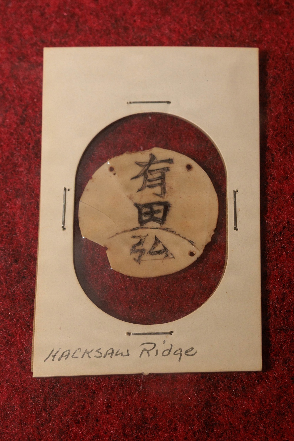 A mounted artifact displayed on a white card with red felt backing, showing a circular disc or tag with Japanese kanji characters written on it. The card is handwritten with the label 'Hacksaw Ridge' at the bottom, indicating this is a war relic from the Battle of Hacksaw Ridge (Okinawa, WWII). The item appears to be a Japanese soldier's identification tag or personal item, pinned to the display card with metal staples or pins.