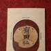 A mounted artifact displayed on a white card with red felt backing, showing a circular disc or tag with Japanese kanji characters written on it. The card is handwritten with the label 'Hacksaw Ridge' at the bottom, indicating this is a war relic from the Battle of Hacksaw Ridge (Okinawa, WWII). The item appears to be a Japanese soldier's identification tag or personal item, pinned to the display card with metal staples or pins.