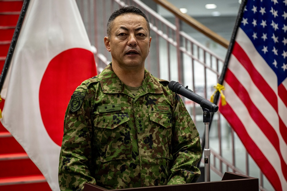 A Japan Ground Self-Defense Force (JGSDF) officer in camouflage uniform is speaking at a podium with a microphone. The setting appears to be an official military ceremony or briefing, flanked by both the Japanese and American flags, suggesting a bilateral military event. The officer's name tag reads 'M. ENDO' and insignia indicates a senior rank.