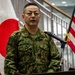 A Japan Ground Self-Defense Force (JGSDF) officer in camouflage uniform is speaking at a podium with a microphone. The setting appears to be an official military ceremony or briefing, flanked by both the Japanese and American flags, suggesting a bilateral military event. The officer's name tag reads 'M. ENDO' and insignia indicates a senior rank.