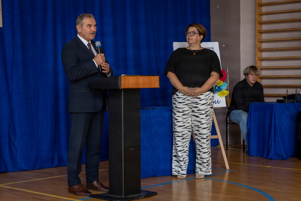 Two adults stand at a podium in what appears to be a school gymnasium during a formal event or presentation. A man in a dark suit is speaking into a microphone at the lectern, while a woman in a black top and zebra-print pants stands beside him. A third person is seated in the background near audio equipment.