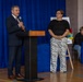 Two adults stand at a podium in what appears to be a school gymnasium during a formal event or presentation. A man in a dark suit is speaking into a microphone at the lectern, while a woman in a black top and zebra-print pants stands beside him. A third person is seated in the background near audio equipment.