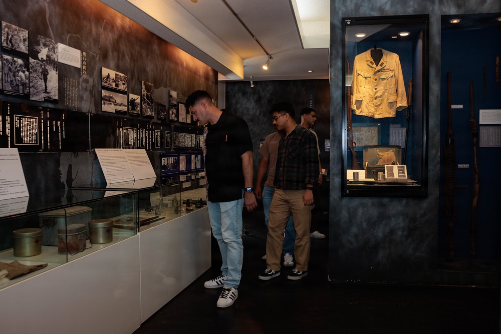 Three visitors are examining exhibits inside what appears to be a war or military history museum. The gallery features black-and-white archival photographs mounted on dark walls, glass display cases containing artifacts, and a mannequin displaying a military uniform jacket in a blue-lit case. The atmosphere is dim and contemplative, typical of a memorial or historical exhibition space.