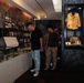 Three visitors are examining exhibits inside what appears to be a war or military history museum. The gallery features black-and-white archival photographs mounted on dark walls, glass display cases containing artifacts, and a mannequin displaying a military uniform jacket in a blue-lit case. The atmosphere is dim and contemplative, typical of a memorial or historical exhibition space.