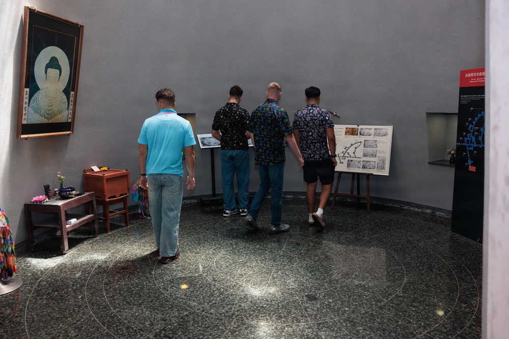 Four men are viewing exhibition displays in what appears to be a museum or memorial hall with Buddhist cultural elements. The space features a Buddha painting on the left wall, informational boards on easels, and a polished granite floor with circular inlay patterns. The setting suggests an educational or commemorative exhibit, likely in an East Asian cultural institution.