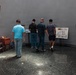 Four men are viewing exhibition displays in what appears to be a museum or memorial hall with Buddhist cultural elements. The space features a Buddha painting on the left wall, informational boards on easels, and a polished granite floor with circular inlay patterns. The setting suggests an educational or commemorative exhibit, likely in an East Asian cultural institution.