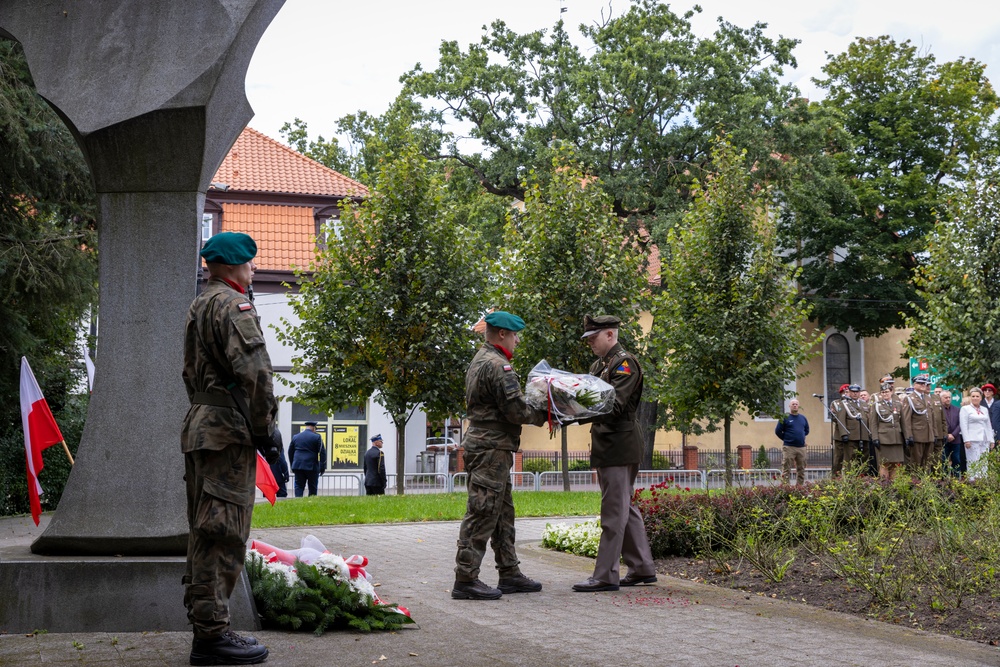 A military wreath-laying ceremony is taking place at a monument, likely in Poland based on the visible Polish flags and uniforms. Soldiers in camouflage uniforms and green berets are transferring a floral wreath near the base of a stone monument. A formation of uniformed personnel and civilians can be seen in the background.