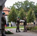 A military wreath-laying ceremony is taking place at a monument, likely in Poland based on the visible Polish flags and uniforms. Soldiers in camouflage uniforms and green berets are transferring a floral wreath near the base of a stone monument. A formation of uniformed personnel and civilians can be seen in the background.