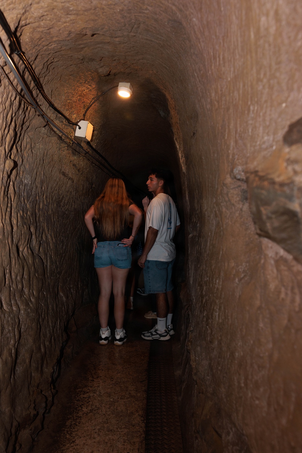 Two young visitors stand inside a narrow, arched stone tunnel that has been fitted with electric lighting and wiring along the ceiling. The tunnel appears to be part of a guided tour attraction, likely a historic mine, cave, or underground passage. One person has turned to look back toward the camera while the other faces forward into the tunnel.