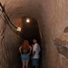 Two young visitors stand inside a narrow, arched stone tunnel that has been fitted with electric lighting and wiring along the ceiling. The tunnel appears to be part of a guided tour attraction, likely a historic mine, cave, or underground passage. One person has turned to look back toward the camera while the other faces forward into the tunnel.