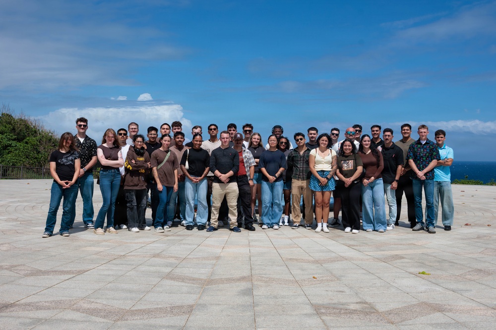 A large group of approximately 30-35 people poses for a formal group photograph on a wide stone-paved plaza or terrace. The setting appears to be an outdoor overlook or memorial site with ocean and blue sky visible in the background. The group appears diverse in age and ethnicity, dressed in casual everyday clothing.