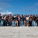 A large group of approximately 30-35 people poses for a formal group photograph on a wide stone-paved plaza or terrace. The setting appears to be an outdoor overlook or memorial site with ocean and blue sky visible in the background. The group appears diverse in age and ethnicity, dressed in casual everyday clothing.