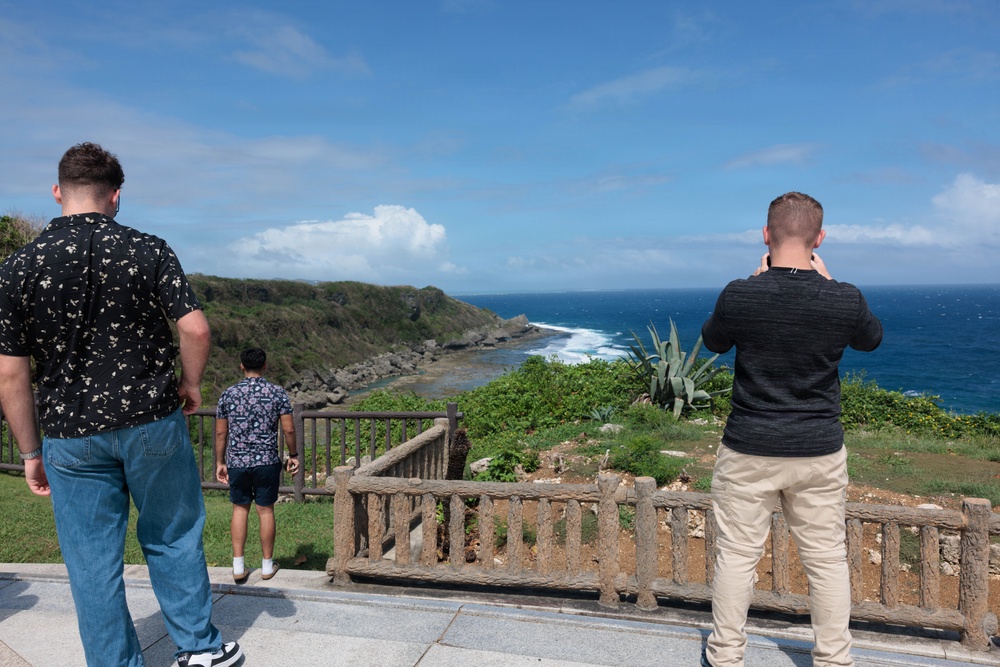 Three individuals stand at a scenic coastal overlook with a rustic wooden railing, gazing at a rocky shoreline and deep blue ocean. The viewpoint appears to be a tourist observation area with lush green vegetation, including agave plants. One person on the right is photographing the landscape with a device held up to their face.