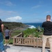 Three individuals stand at a scenic coastal overlook with a rustic wooden railing, gazing at a rocky shoreline and deep blue ocean. The viewpoint appears to be a tourist observation area with lush green vegetation, including agave plants. One person on the right is photographing the landscape with a device held up to their face.