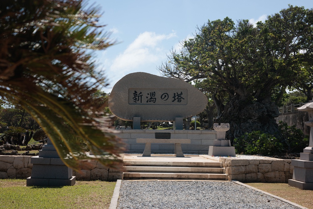The image shows a Japanese memorial monument with the inscription '新潟の塔' (Niigata no To / Tower of Niigata), located in what appears to be a war memorial park, likely in Okinawa, Japan. The stone monument sits on a raised platform accessed by steps, surrounded by mature trees and manicured grounds. This type of memorial is characteristic of the Peace Memorial Park in Mabuni, Okinawa, where prefectures erected monuments to honor their war dead.