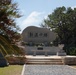 The image shows a Japanese memorial monument with the inscription '新潟の塔' (Niigata no To / Tower of Niigata), located in what appears to be a war memorial park, likely in Okinawa, Japan. The stone monument sits on a raised platform accessed by steps, surrounded by mature trees and manicured grounds. This type of memorial is characteristic of the Peace Memorial Park in Mabuni, Okinawa, where prefectures erected monuments to honor their war dead.