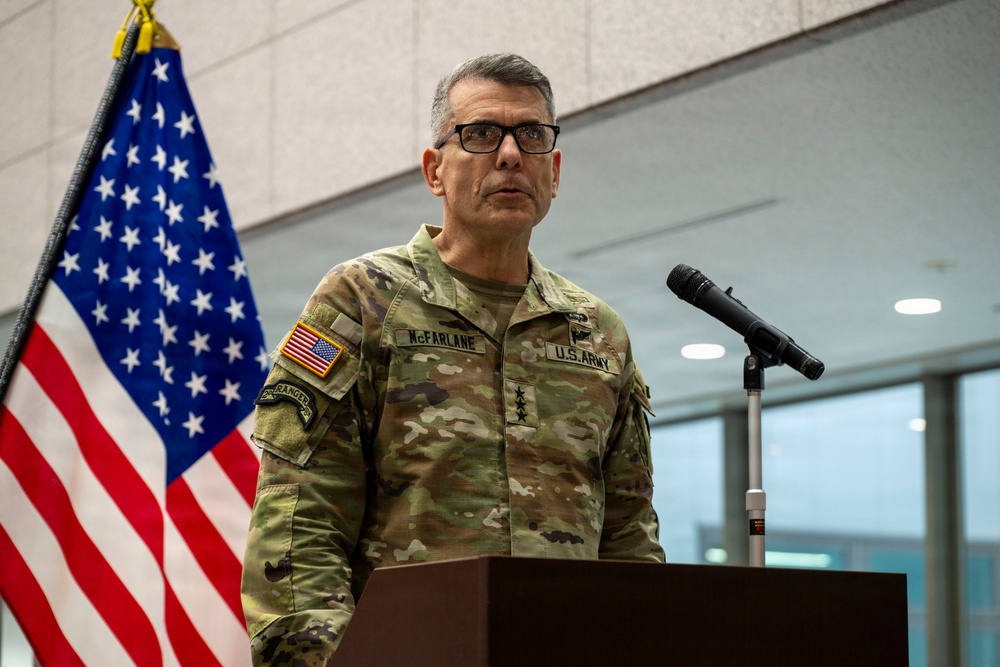 A U.S. Army Lieutenant General named McFarlane is speaking at a podium with a microphone. He is wearing MultiCam (OCP) camouflage uniform with three-star rank insignia visible on his chest. An American flag is displayed behind him to the left.