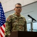 A U.S. Army Lieutenant General named McFarlane is speaking at a podium with a microphone. He is wearing MultiCam (OCP) camouflage uniform with three-star rank insignia visible on his chest. An American flag is displayed behind him to the left.