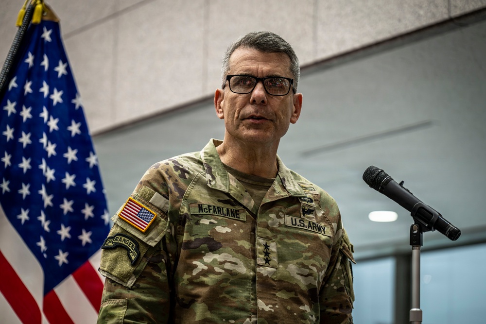 A U.S. Army lieutenant general named McFarlane is speaking at a podium with a microphone. He is wearing an OCP (Operational Camouflage Pattern) uniform with visible name tape, U.S. Army tape, a Ranger tab, and three-star rank insignia. An American flag is visible in the background.