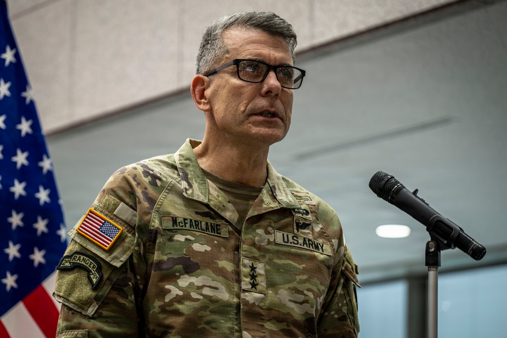 A U.S. Army Lieutenant General named McFarlane is speaking at a podium with a microphone. He is wearing an OCP (Operational Camouflage Pattern) uniform with three-star rank insignia visible on his chest. A U.S. flag is partially visible in the background to his left.