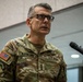 A U.S. Army Lieutenant General named McFarlane is speaking at a podium with a microphone. He is wearing an OCP (Operational Camouflage Pattern) uniform with three-star rank insignia visible on his chest. A U.S. flag is partially visible in the background to his left.