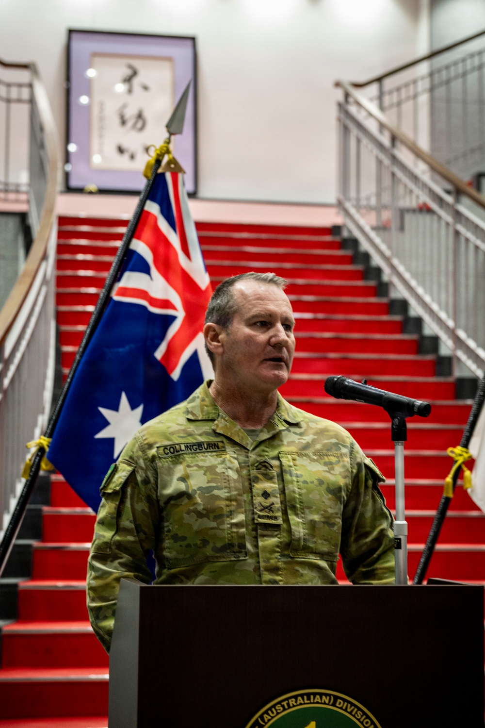 An Australian Army officer named Collingburn is speaking at a podium during an official military ceremony or briefing. He is wearing Australian Multicam Camouflage (AUSCAM/Multicam) uniform with rank insignia visible on his chest. The setting appears to be an indoor venue with a red-carpeted staircase and the Australian national flag displayed behind him.