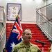 An Australian Army officer named Collingburn is speaking at a podium during an official military ceremony or briefing. He is wearing Australian Multicam Camouflage (AUSCAM/Multicam) uniform with rank insignia visible on his chest. The setting appears to be an indoor venue with a red-carpeted staircase and the Australian national flag displayed behind him.