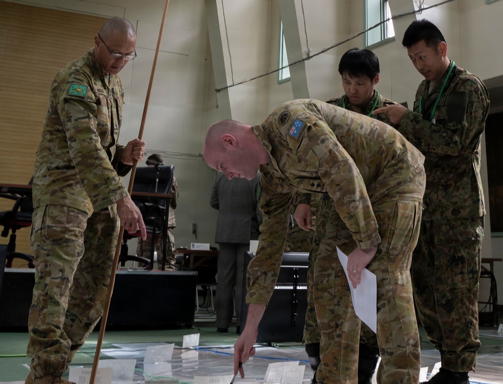 Four military personnel in camouflage uniforms are gathered around a large floor map or tactical display in what appears to be an indoor briefing or planning session. Two individuals wearing Australian Army patches are actively pointing at and examining the map, while two others in different uniforms observe. A civilian in a suit is visible in the background.