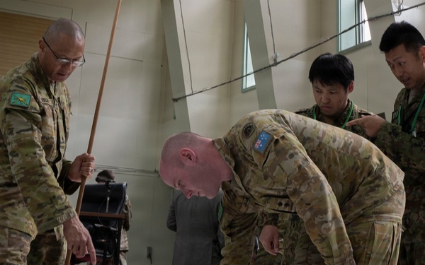 Four military personnel in camouflage uniforms are gathered around a large floor map or tactical display in what appears to be an indoor briefing or planning session. Two individuals wearing Australian Army patches are actively pointing at and examining the map, while two others in different uniforms observe. A civilian in a suit is visible in the background.