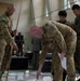 Four military personnel in camouflage uniforms are gathered around a large floor map or tactical display in what appears to be an indoor briefing or planning session. Two individuals wearing Australian Army patches are actively pointing at and examining the map, while two others in different uniforms observe. A civilian in a suit is visible in the background.